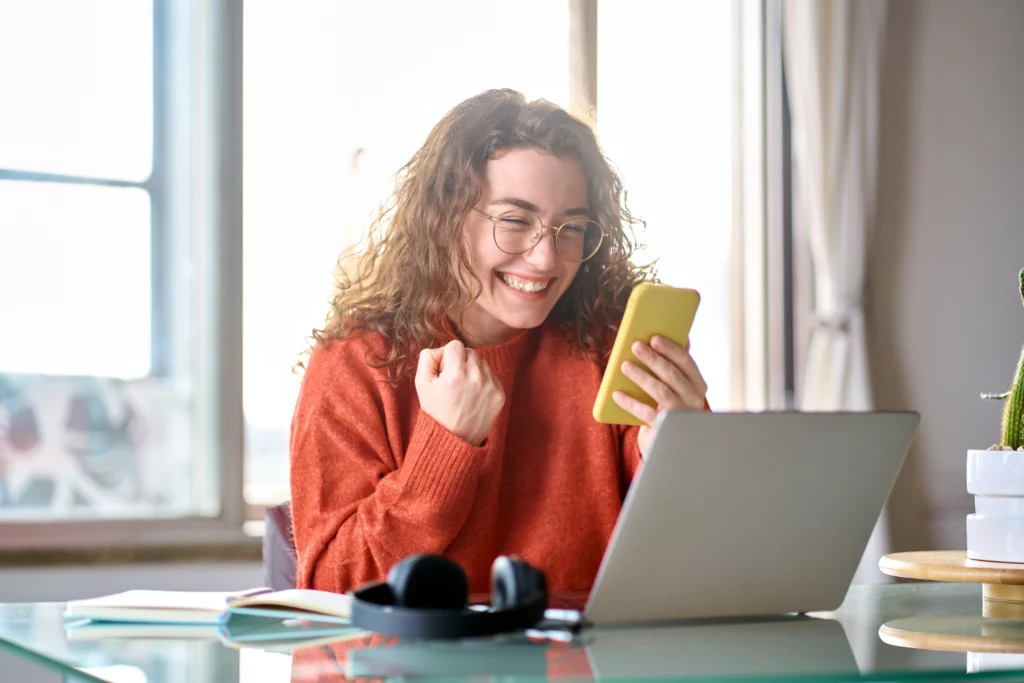 A young woman on her phone sitting in front of a laptop celebrates the approval of a loan